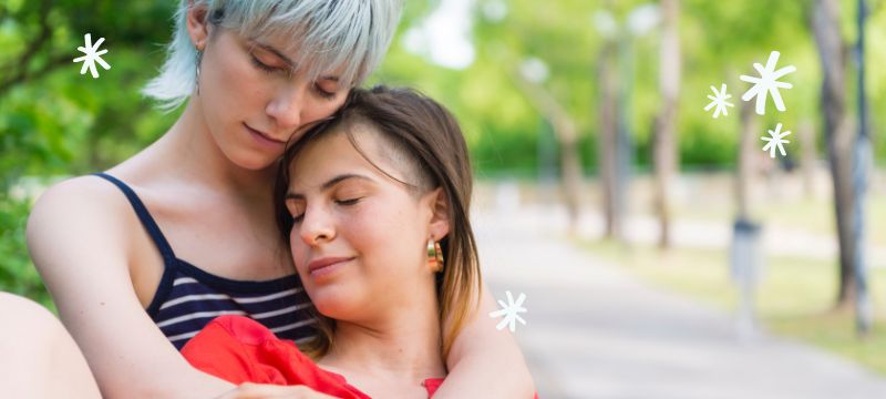 A woman with short silver hair sits behind a woman in a red top hugging her. They both have their eyes closed